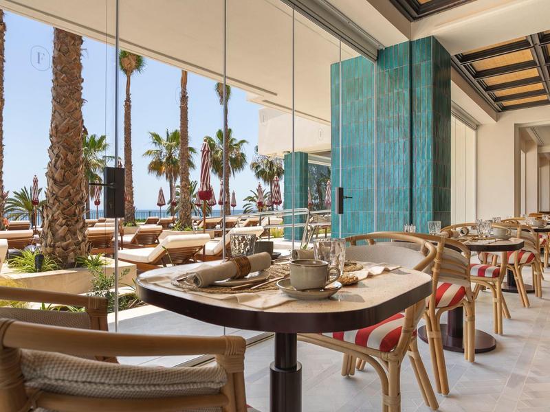 Restaurantterrasse mit Holztischen, Stühlen, Palmen und Blick auf die Küste bei Sonnenschein.