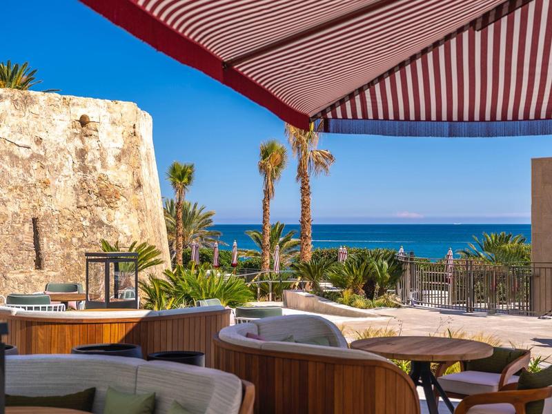 Strandterrasse mit Tischen, Stühlen und Sonnenschirm, Blick auf Meer und Felsen bei Sonnenschein.