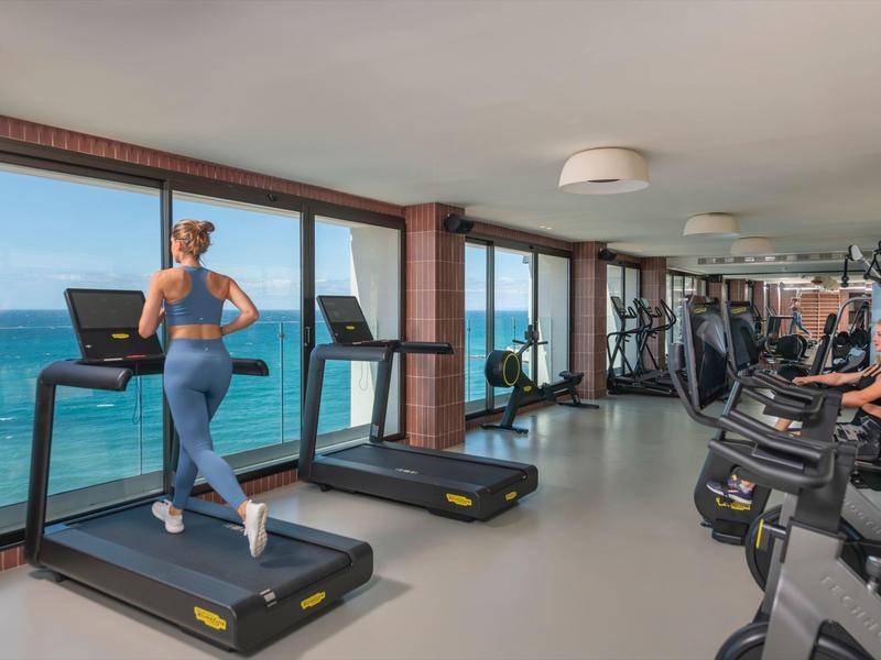 Fitnessraum mit großen Fenstern, Laufbändern, Trainingsrädern und Blick auf das Meer.
