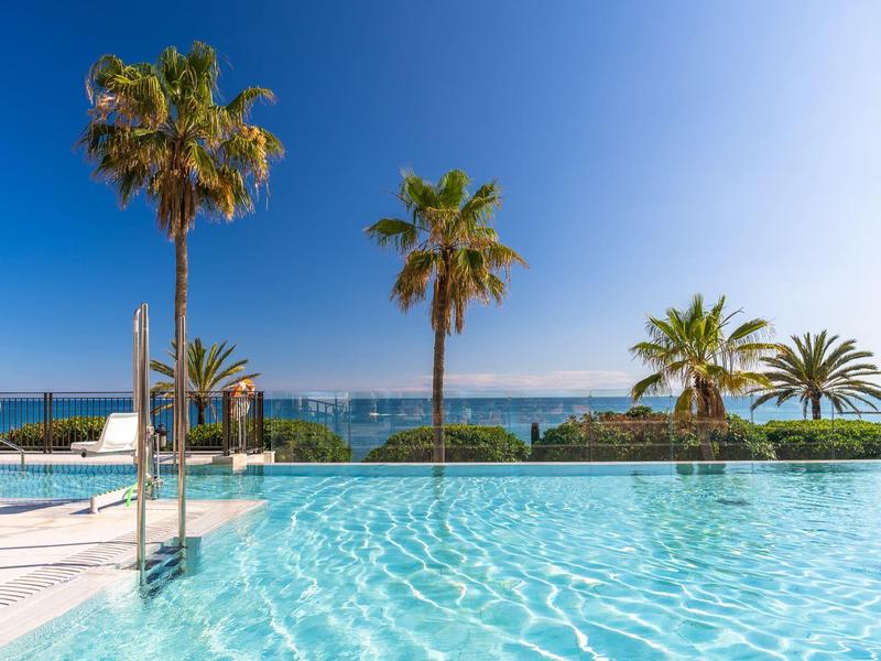 Ein klarer blauer Pool mit Palm Bäumen vor dem ruhigen blauen Meer unter klarem Himmel.