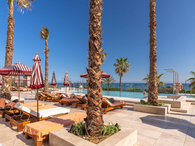 Strandpromenade mit Palmen, Liegestühlen und Sonnenschirmen am Meer bei klarem Himmel.