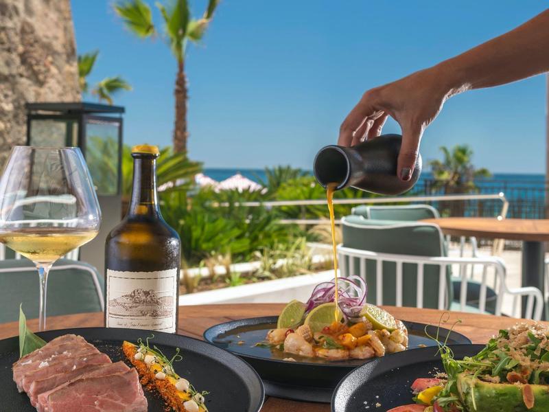 Sommerliche Terrasse mit gedecktem Tisch, Weinflasche, Glas und Hand gießt Soße auf Essen.
