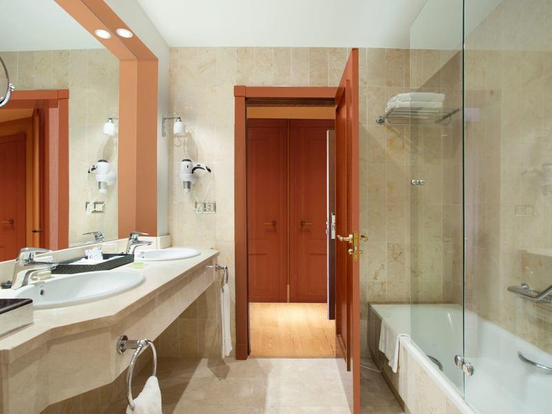 Helles Badezimmer mit großer Spiegelwand, Doppelwaschbecken, Badewanne und Holztür.