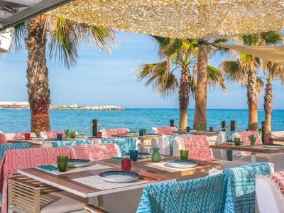 Terrasse eines Strandrestaurants mit bunten Sitzkissen und Blick auf das Meer und Palmen.