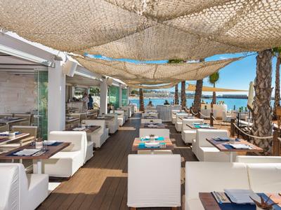 Offenes Strandrestaurant mit weißen Sitzmöbeln und Blick auf das blaue Meer unter Sonnensegeln.