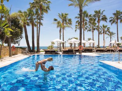 Piscine d'hôtel avec palmiers et parasols sur la plage sous un ciel clair.