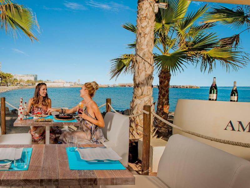 Zwei Frauen sitzen an einem gedeckten Tisch im Freien am Strand unter Palmen.