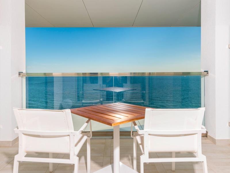Balkon mit zwei weißen Stühlen und kleinem Holztisch mit Blick auf das Meer unter klarem Himmel.
