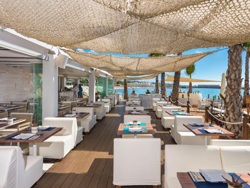 Offenes Strandrestaurant mit weißen Sitzmöbeln und Blick auf das blaue Meer unter Sonnensegeln.