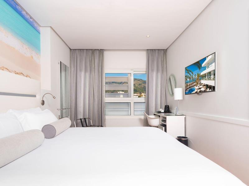 Helles Hotelzimmer mit großem Bett, Wandbild und Fenster mit Blick auf das Wasser.