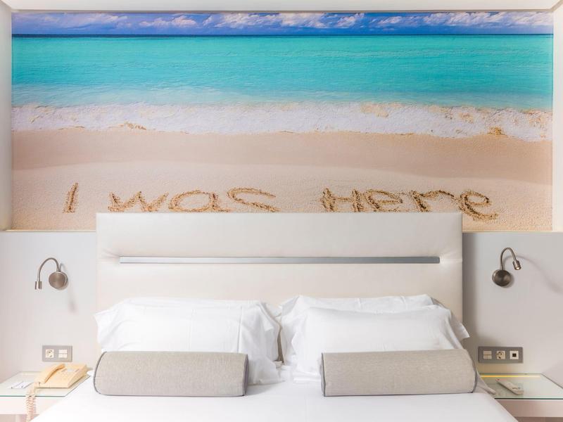 Modernes Hotelzimmer mit weißem Bett und Wandbild eines Strandes mit der Aufschrift "I was here" im Sand.