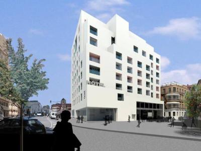 Modernes, weißes Gebäude mit großen Fenstern an einer breiten Straßenecke, Bäume und Menschen.