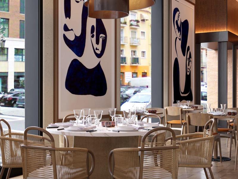 Restaurant mit rundem Tisch, Weingläsern und Stühlen aus geflochtenem Holz vor großen Fenstern.