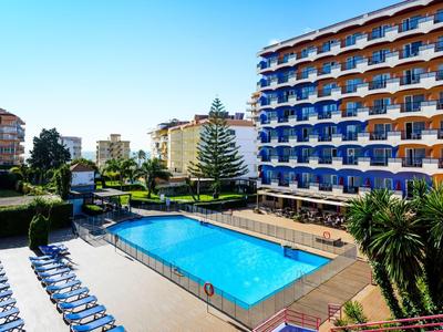 Großer Außenpool vor einem mehrstöckigen Hotel mit blauen Balkonen und Sonnenliegen.