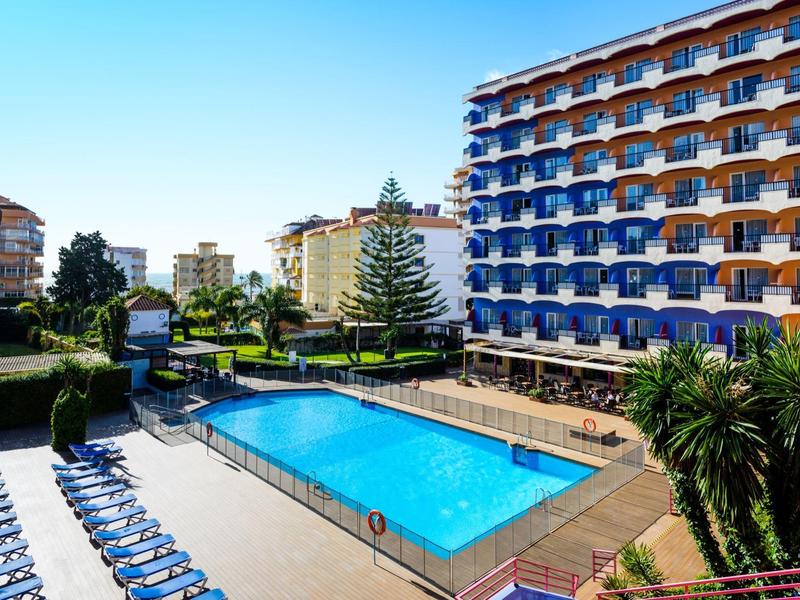 Großer Außenpool vor einem mehrstöckigen Hotel mit blauen Balkonen und Sonnenliegen.