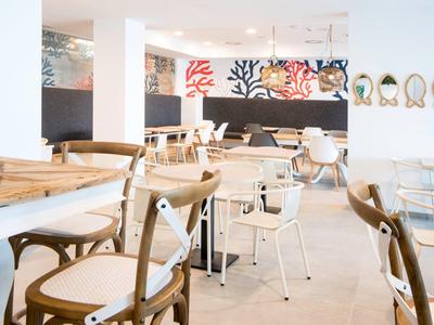 Helles Café mit Holz- und Metallstühlen, weißen Tischen und bunten Wandbildern im Hintergrund.