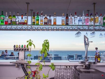 Bar mit bunten Spirituosenflaschen, Glasregalen und Blick auf Meer und bewölkten Himmel.