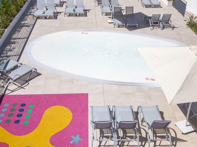 Leerer Poolbereich mit weißen Liegestühlen, buntem Bodenmuster und Spielplatz im Hintergrund.