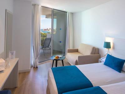 Helles Hotelzimmer mit zwei Betten, Balkon, Sofa, Tisch, Fernseher und weiß-blauer Farbkombination.