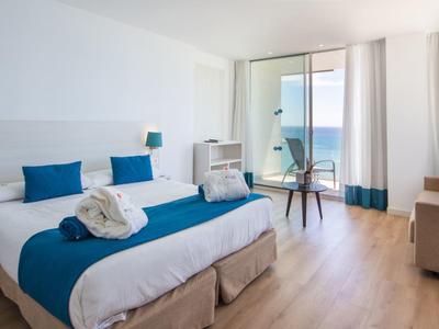 Helles Hotelzimmer mit Doppelbett, blauen Akzenten und Meerblick durch große Fenster.