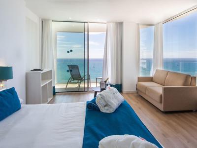 Moderner Hotelraum mit großem Bett, Sofa, Holzboden und Balkon mit Meerblick.