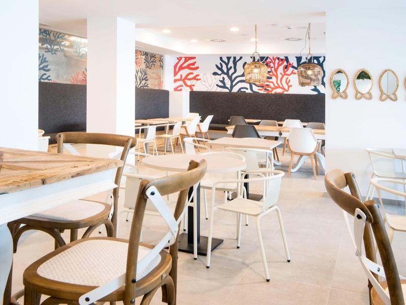 Helles Café mit Holz- und Metallstühlen, weißen Tischen und bunten Wandbildern im Hintergrund.