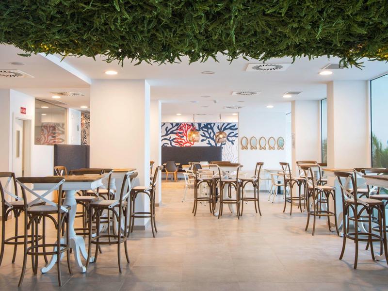 Moderner, heller Café-Raum mit Holzstühlen, Tischen und grüner Deckenpflanzen-Dekoration.