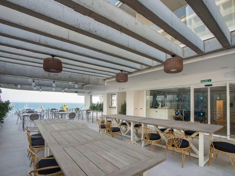 Überdachte Terrasse mit langen Holztischen, geflochtenen Stühlen und Blick auf das Meer.