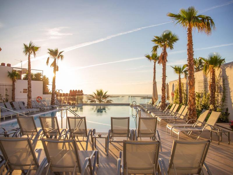 Sonnige Dachterrasse mit Pool, Liegestühlen und Palmen, Meerblick im Hintergrund bei klarem Himmel.