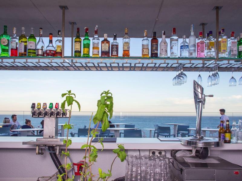 Bar mit bunten Spirituosenflaschen, Glasregalen und Blick auf Meer und bewölkten Himmel.