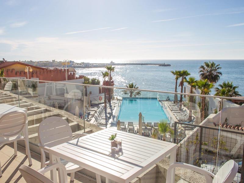 Terrasse mit weißen Stühlen und Tischen, Blick auf Pool, Palmen und blaues Meer bei klarem Himmel.
