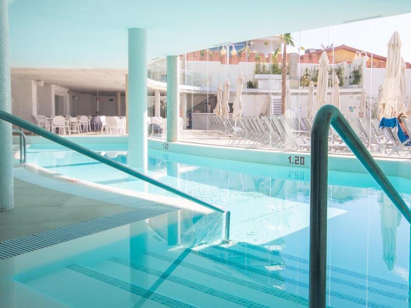 Moderner Innenpool mit klar blauer Wasserfläche, Treppen und Edelstahlgeländern. Helle Wände und große Fenster.