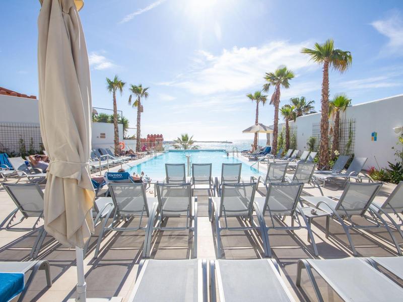 Moderner Poolbereich mit Liegestühlen, Sonnenschirmen und Palmen unter klarem, blauem Himmel.