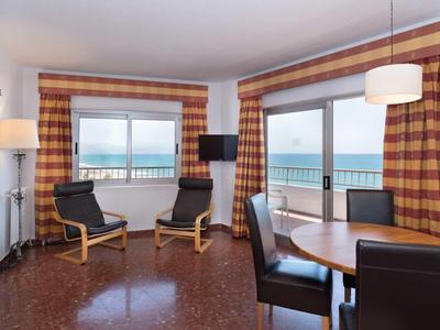 Helles Zimmer mit großen Fenstern, zwei Stühlen, Glastisch, braun karierten Vorhängen und Meeresblick.