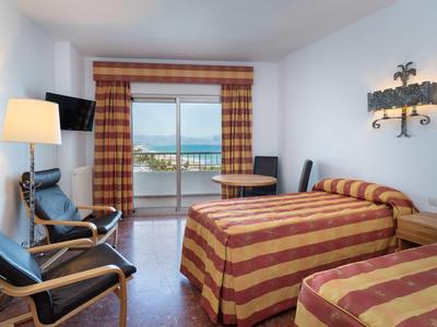 Helles Hotelzimmer mit zwei Betten, Sesseln, Fernseher und Balkon mit Meeresblick.