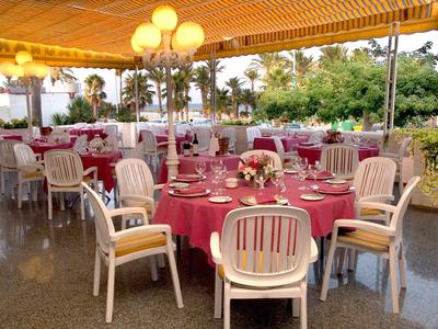 Restaurantterrasse mit rund gedeckten Tischen, roten Tischdecken und weißen Holzstühlen unter Holzdecke.