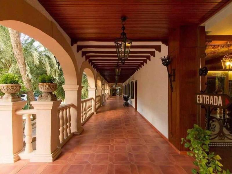 Langer Flur mit dunkelrotem Fliesenboden, Holzdach und Säulenbogen, grünes Pflanzenarrangement links.