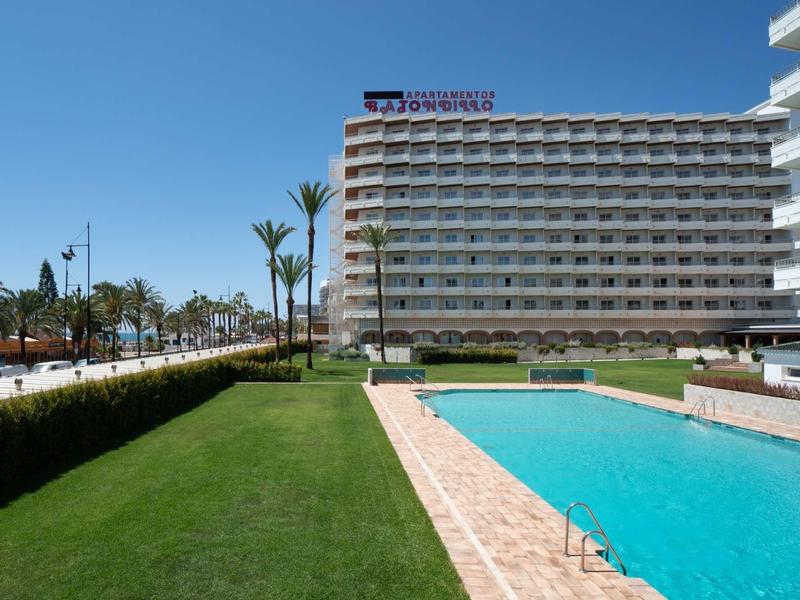 Großes Hotel mit mehreren Stockwerken neben einem Pool und gepflegtem Rasen unter blauem Himmel.