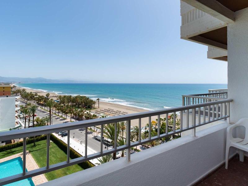 Balkon mit weißen Stühlen und Geländer, Blick auf Pool, Palmen, Strand und blaues Meer unter klarem Himmel.