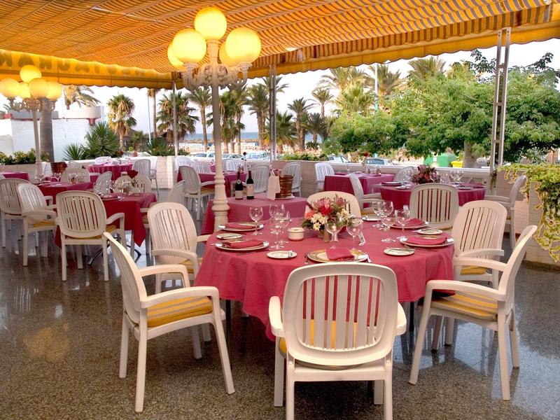 Restaurantterrasse mit rund gedeckten Tischen, roten Tischdecken und weißen Holzstühlen unter Holzdecke.