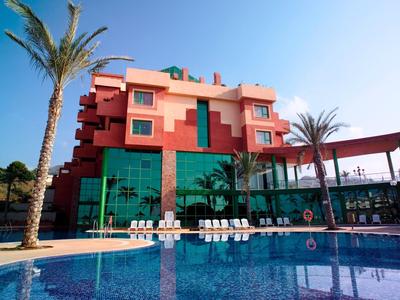 Innenraum mit grauem Metalltisch, buntem Teppich und gelber Stehlampe vor roter Ziegelwand.