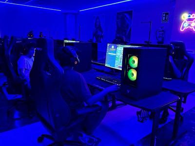 Dunkler Raum mit blauer Beleuchtung, zwei Personen an PCs mit Spiele-Setup und Neon-Gaming-Zeichen.