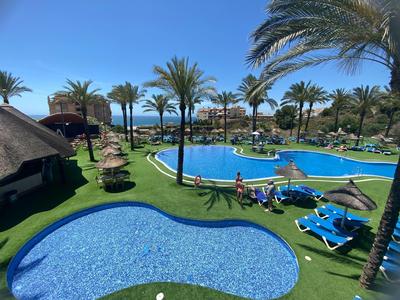 Poollandschaft in einem Hotel mit Palmen, Liegestühlen und Sonnenschirmen unter blauem Himmel.