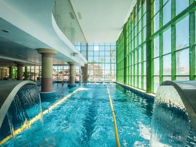 Brightly lit indoor pool with waterfalls and large green windows.