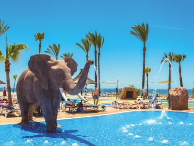 Pool with fountain and elephant statue beside palm trees and sun loungers under clear sky