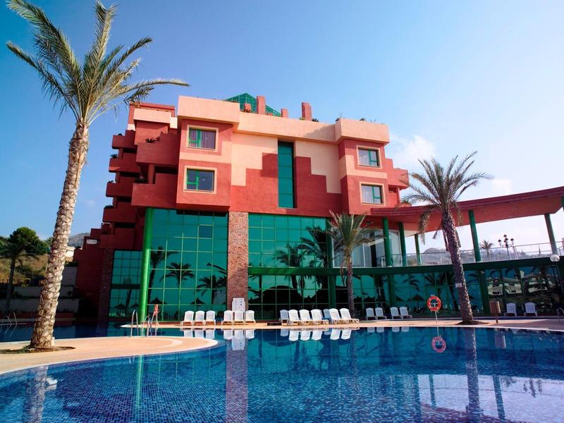 Innenraum mit grauem Metalltisch, buntem Teppich und gelber Stehlampe vor roter Ziegelwand.