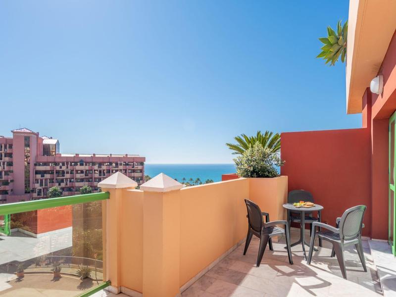 Balkon mit Tisch und Stühlen, Blick auf Meer und Gebäude unter klarem Himmel.