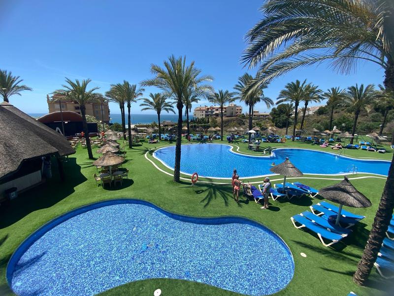 Poollandschaft in einem Hotel mit Palmen, Liegestühlen und Sonnenschirmen unter blauem Himmel.