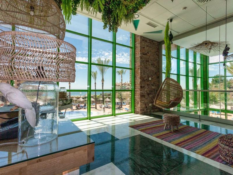 Modernes, sonnendurchflutetes Zimmer mit großen grünen Fenstern und bunten Teppichen in einer Hotelanlage.