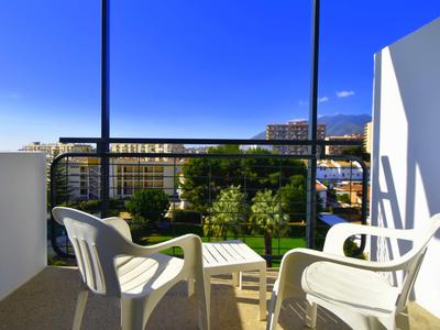 Balkon mit Blick auf einen Garten und Gebäude, zwei weiße Stühle und ein kleiner Tisch.